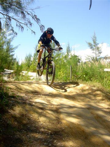 A mountain biker in mid-air over a dirt ramp, wearing a helmet and riding gear, surrounded by greenery and trees under a clear blue sky. Oleta River State Park mountain bike trail.