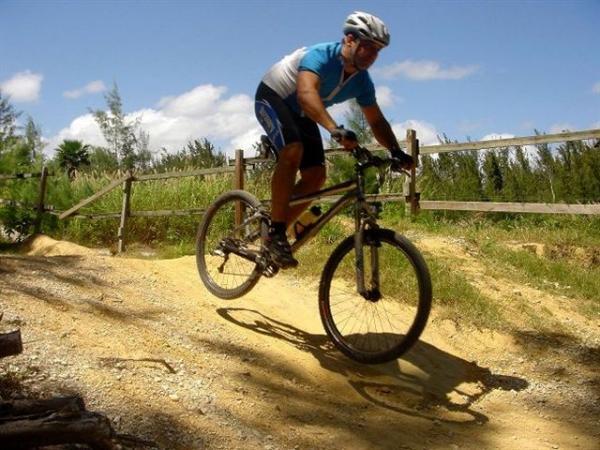 A mountain biker in a blue and white jersey rides over a dirt path, catching air as he jumps off a small hill. The background features a wooden fence and greenery under a blue sky with scattered clouds. Oleta River State Park mountain bike trail.