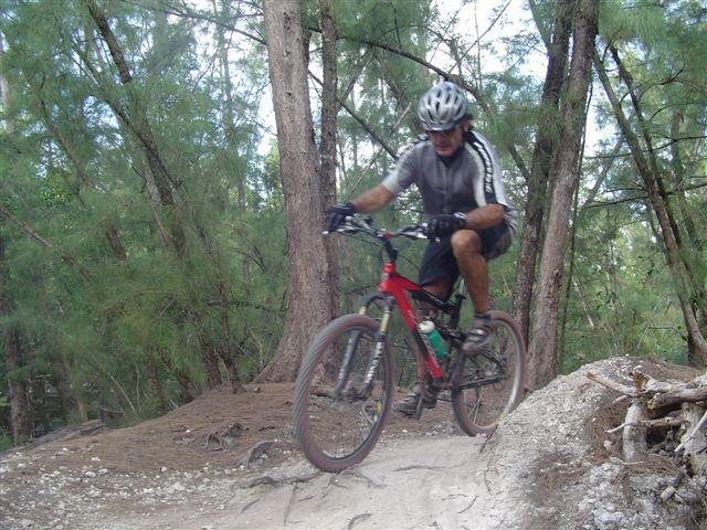 A mountain biker riding on a dirt trail in a wooded area, surrounded by tall trees. The cyclist is wearing a helmet and cycling gear, navigating a rocky section of the path with one wheel lifted off the ground. Oleta River State Park mountain bike trail.