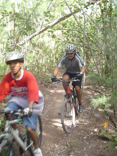 Two cyclists navigate a dirt trail in a wooded area. One cyclist, wearing a red shirt and a helmet, is in the foreground, while a second cyclist, dressed in a gray outfit and also wearing a helmet, is positioned slightly behind. Lush greenery surrounds the trail, creating a natural backdrop for their ride. Oleta River State Park mountain bike trail.