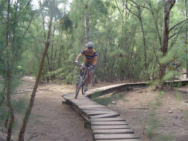 A mountain biker navigating a wooden plank trail through a dense forest. The cyclist is wearing a helmet and a colorful cycling jersey, with one wheel lifted as they maneuver along the winding path surrounded by trees. Oleta River State Park mountain bike trail.