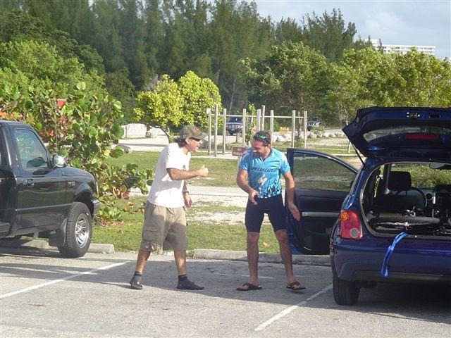 Two men are interacting outdoors in a parking lot, surrounded by trees. One man, wearing a white t-shirt and khaki shorts, appears to be gesturing animatedly, while the other man is dressed in a blue athletic shirt and black shorts, smiling as he responds. Their vehicles are parked nearby, and the scene conveys a sense of casual camaraderie. Oleta River State Park mountain bike trail.