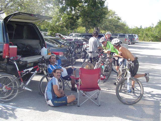 A group of cyclists resting near their parked vehicles in a lot. Some are seated on folding chairs, while others are preparing their bicycles. The scene is sunny, with trees in the background and various cars present. Oleta River State Park mountain bike trail.