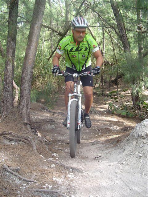 A mountain biker dressed in a bright green jersey and black shorts navigates a dirt trail surrounded by trees. The cyclist is mid-jump, showcasing an action-packed moment in a forested area. Oleta River State Park mountain bike trail.