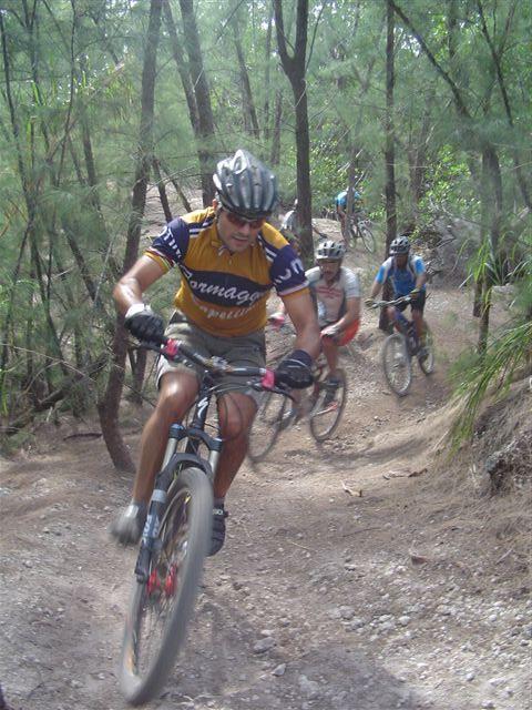 A group of mountain bikers navigating a rugged, tree-lined trail. The lead cyclist, wearing a yellow jersey with blue accents and a helmet, is in an active riding position. Two other cyclists follow closely behind on the winding path. Sunlight filters through the trees, illuminating the natural surroundings. Oleta River State Park mountain bike trail.