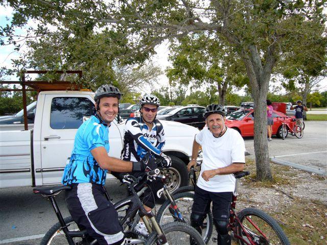 Three mountain bikers wearing helmets and cycling attire smile at the camera while posed next to their bikes in a parking area surrounded by trees. A pickup truck and several cars are visible in the background, suggesting an outdoor biking location. Oleta River State Park mountain bike trail.