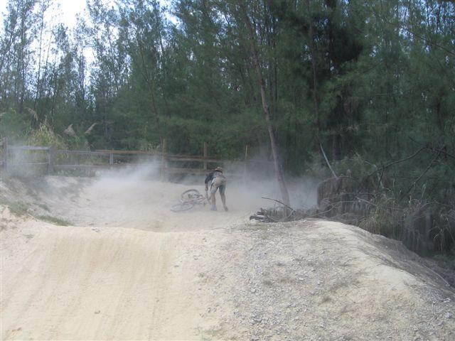 A mountain biker falls off their bike on a dirt trail, surrounded by trees. Dust clouds are visible in the air, indicating recent movement. Oleta River State Park mountain bike trail.