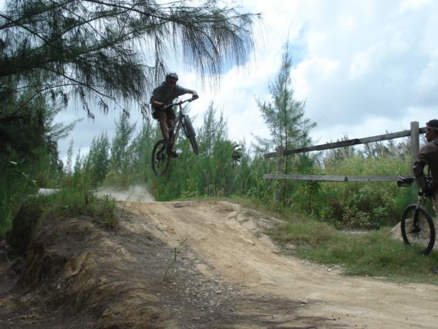 A cyclist performing a jump on a mountain bike off a dirt ramp surrounded by greenery and trees on a sunny day. Another cyclist can be seen in the background, observing the jump. Dust is kicked up from the ramp as the rider launches into the air. Oleta River State Park mountain bike trail.