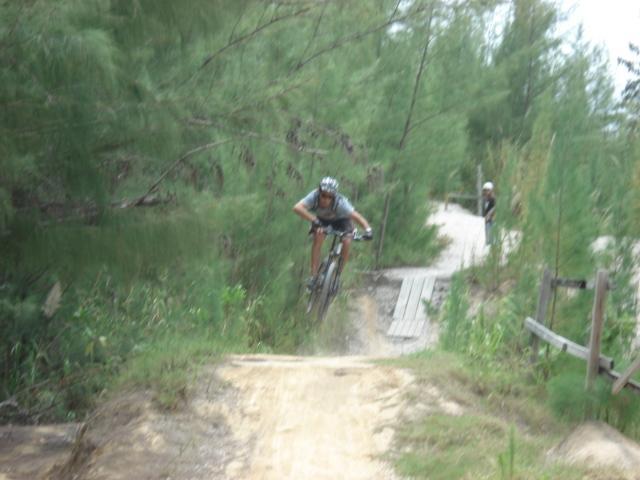 A mountain biker jumps over a dirt ramp on a wooded trail, surrounded by tall trees. In the background, a second person watches from a distance. The scene captures the thrill of biking in a natural setting. Oleta River State Park mountain bike trail.