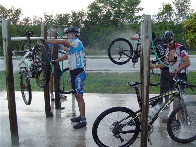 A group of cyclists washing their mountain bikes at a bike cleaning station. Two cyclists are actively spraying their bikes with water, while a third cyclist prepares to clean another bike. The setting appears to be outdoors, surrounded by greenery, with a road visible in the background. Oleta River State Park mountain bike trail.