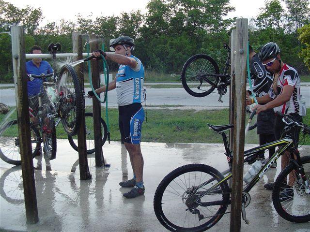 Three cyclists are cleaning their mountain bikes at a bike wash station. One cyclist is holding a bike on a rack and using a hose to wash it, while another cyclist stands nearby, rinsing off their bike. The scene is set in an outdoor area with greenery in the background, and the sun is shining. Oleta River State Park mountain bike trail.