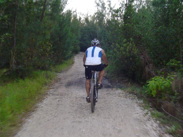 A person riding a bicycle on a gravel path surrounded by trees and greenery, viewed from the back. The cyclist is wearing a white sleeveless top with blue stripes and black shorts. Oleta River State Park mountain bike trail.