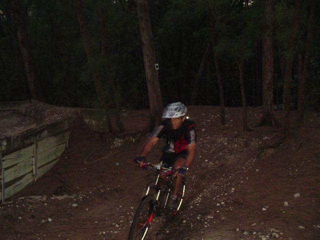 A mountain biker wearing a helmet and cycling gear navigates a dirt trail in a forested area during dusk. The terrain is rough, with patches of dirt and gravel, and tall trees surround the path. Oleta River State Park mountain bike trail.