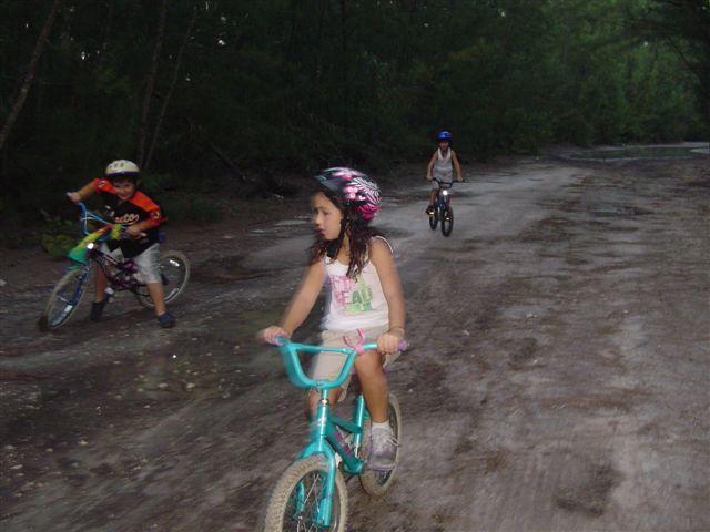 Three children riding bicycles on a dirt road surrounded by greenery. One child is wearing a pink helmet and riding a light blue bicycle, while another child in an orange shirt is riding a bike in the background. The third child is also on a bike further down the path. The scene captures a fun outdoor activity in a natural setting. Oleta River State Park mountain bike trail.