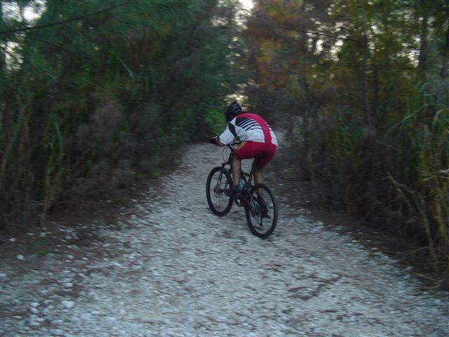 A person riding a mountain bike along a gravel path surrounded by dense vegetation in a wooded area. The rider is dressed in a black and red cycling outfit and is seen from behind, navigating the trail. Oleta River State Park mountain bike trail.