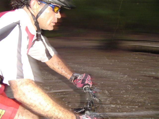 A cyclist in motion on a muddy trail, wearing a helmet, sunglasses, and a cycling jersey. The image captures the dynamic movement, with the background blurred to emphasize speed. The cyclist has mud splattered on his arms and gear, suggesting an adventurous ride through rough terrain. Oleta River State Park mountain bike trail.