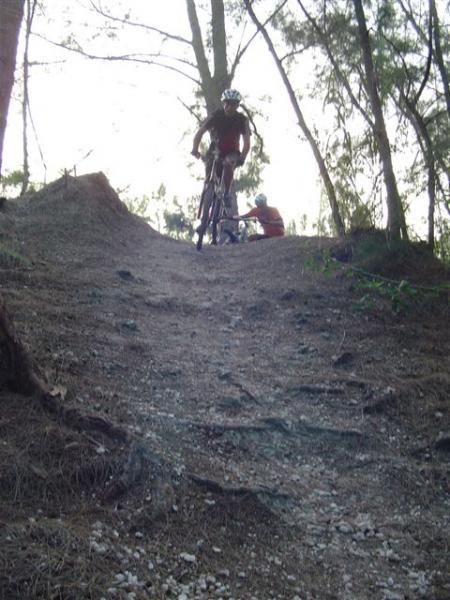 Mountain biker ascending a dirt hill in a forested area, with another person visible in the background. The scene captures the natural surroundings with trees and earthy terrain. Oleta River State Park mountain bike trail.
