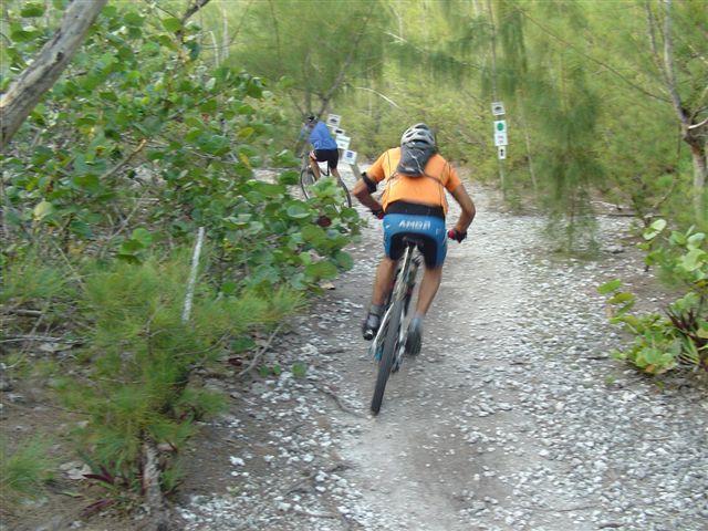 A person riding a mountain bike down a rocky trail surrounded by greenery. The cyclist is wearing a helmet and an orange shirt, with another cyclist visible in the background. Trail markers can be seen along the path. Oleta River State Park mountain bike trail.
