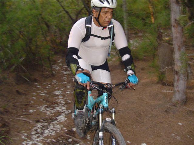 A person riding a mountain bike on a narrow dirt trail surrounded by trees. The cyclist is wearing a helmet, padded clothing, and protective gear, demonstrating an active outdoor sports activity. Oleta River State Park mountain bike trail.