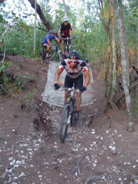 A group of three mountain bikers navigate a rugged trail in a forested area. One rider, in the foreground, is descending a steep, rough section of the path, showcasing balance and skill. The other two riders are slightly behind, positioned on a less steep part of the terrain, surrounded by lush greenery and trees. The ground is uneven, with scattered rocks and dirt, highlighting the adventurous nature of the sport. Oleta River State Park mountain bike trail.