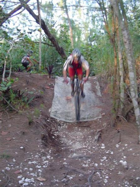 A mountain biker in red shorts and a helmet is airborne while jumping off a dirt ramp in a wooded trail. In the background, another cyclist is positioned on the trail. The scene captures the excitement of mountain biking in a natural setting with greenery and dirt paths. Oleta River State Park mountain bike trail.
