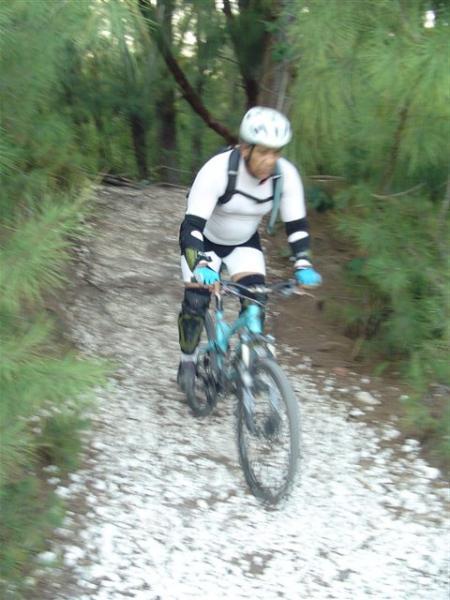 A blurred image of a person biking on a gravel trail surrounded by lush greenery. The cyclist is wearing a helmet and protective gear, and is focused on navigating the path. Oleta River State Park mountain bike trail.