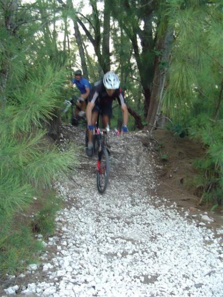 Two mountain bikers navigating a rocky trail through a forested area. One rider is in the foreground, airborne as they navigate a section of loose gravel, while the other is visible in the background. The surrounding vegetation consists of tall trees and greenery, creating a natural setting. Oleta River State Park mountain bike trail.
