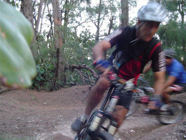 A blurred image of two mountain bikers navigating a dirt trail surrounded by trees. One biker is in the foreground, wearing a black and red cycling jersey, while the other is slightly behind, dressed in a blue shirt. The motion suggests an action-packed biking experience in a natural environment. Oleta River State Park mountain bike trail.