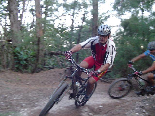 A blurred action shot of two mountain bikers navigating a dirt trail surrounded by trees. One rider is in the foreground, wearing a red and white jersey with a helmet, leaning into a turn, while the second rider, slightly behind, is in a blue jersey. The scene conveys speed and agility in a natural outdoor setting. Oleta River State Park mountain bike trail.
