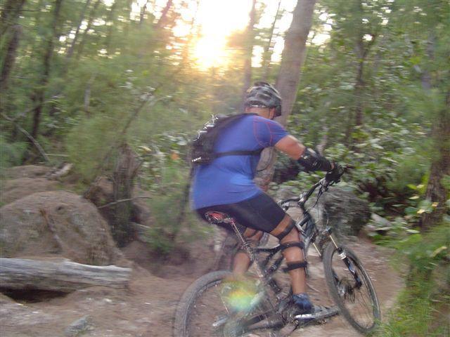 A cyclist wearing a blue shirt and a helmet rides a mountain bike along a dirt trail in a wooded area, with sunlight filtering through the trees in the background. Oleta River State Park mountain bike trail.
