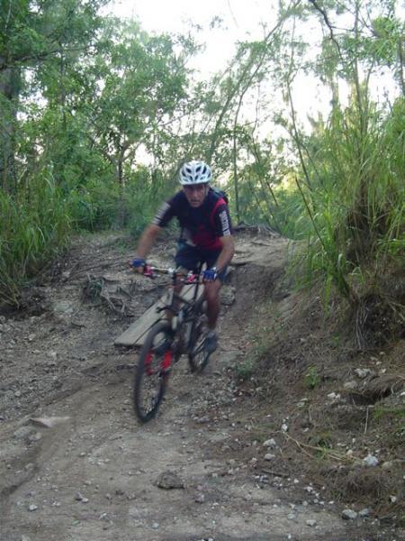 A mountain biker wearing a helmet and cycling gear rides a bike on a dirt trail surrounded by greenery and trees. The rider is navigating a rough section of the path that includes a small wooden ramp. Oleta River State Park mountain bike trail.