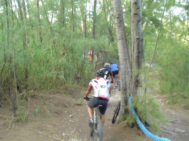 A group of mountain bikers navigating a dirt trail through a wooded area. The path is surrounded by greenery and trees, with one biker in the foreground wearing a white and red jersey and another in a blue shirt following closely behind. Oleta River State Park mountain bike trail.