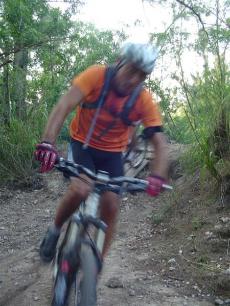 A blurry photograph of a mountain biker in motion, wearing an orange shirt and a helmet, riding on a dirt trail surrounded by greenery. The image captures the dynamic movement of the cyclist as they navigate the rough terrain. Oleta River State Park mountain bike trail.