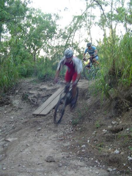 Two mountain bikers navigating a dirt trail, with one cyclist in a red outfit focused on crossing a wooden plank bridge while the other cyclist follows in the background. Surrounding vegetation includes tall grass and trees, indicating a natural outdoor setting. Oleta River State Park mountain bike trail.