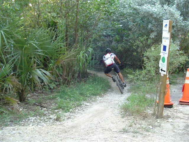 A person riding a mountain bike along a dirt trail surrounded by lush greenery. The cyclist is seen from behind, navigating a curve in the path. There are traffic cones and a directional sign indicating trail routes nearby. Oleta River State Park mountain bike trail.