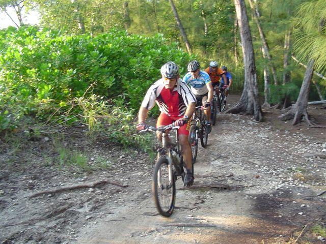 A group of four mountain bikers riding along a dirt trail surrounded by greenery and trees, with the lead rider wearing a red and white jersey. The scene captures an outdoor adventure in a natural setting. Oleta River State Park mountain bike trail.