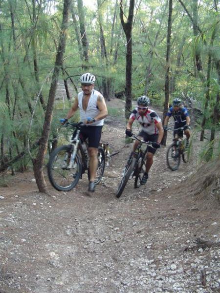 A group of three mountain bikers navigating a gravel path in a wooded area. The cyclists are wearing helmets and cycling gear, with one biker pushing his bike up a hill while the others ride behind him. The background features tall trees and greenery, creating a natural, outdoor setting. Oleta River State Park mountain bike trail.