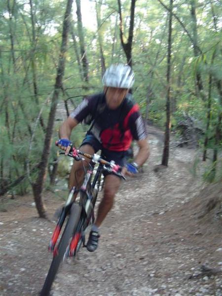 A cyclist wearing a helmet and racing gear is navigating a dirt trail through a wooded area, leaning forward as they ride swiftly along a winding path surrounded by trees. Oleta River State Park mountain bike trail.