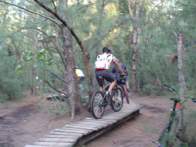 Two mountain bikers riding along a wooden planked trail through a forested area, surrounded by trees and greenery. One biker is wearing a white and red jersey, while the other is in a darker outfit. The scene captures the essence of outdoor recreation and cycling. Oleta River State Park mountain bike trail.