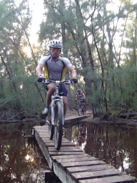 Mountain bikers riding on a narrow wooden bridge over water, surrounded by lush trees and foliage. The image captures one cyclist in the foreground, focused on maintaining balance, while another cyclist is seen in the background. The setting conveys an adventurous and picturesque outdoor scene. Oleta River State Park mountain bike trail.