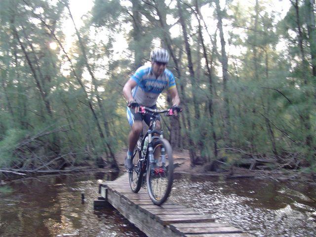 A mountain biker riding across a narrow wooden bridge over a calm body of water, surrounded by trees. The cyclist is wearing a blue and white jersey and a helmet, showcasing a focused expression as they navigate the challenging terrain. Oleta River State Park mountain bike trail.