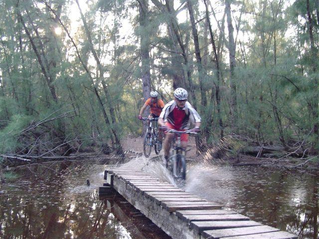 Two mountain bikers navigating a wooden bridge over a waterlogged trail in a forested area. The first rider, wearing an orange shirt and helmet, appears to be further back while the second rider, donning a red and white jersey, is launching off the bridge, splashing water as they pedal. Lush greenery surrounds the scene, with soft sunlight filtering through the trees. Oleta River State Park mountain bike trail.