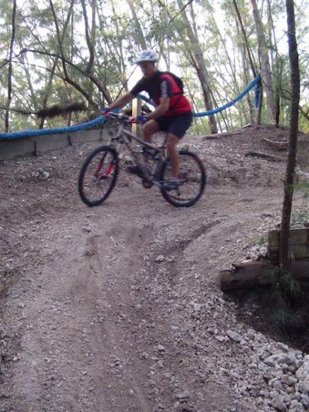 A mountain biker in mid-air, performing a jump on a dirt trail surrounded by trees. The cyclist is wearing a helmet and a red and black jersey, showcasing an active and adventurous outdoor scene. Oleta River State Park mountain bike trail.