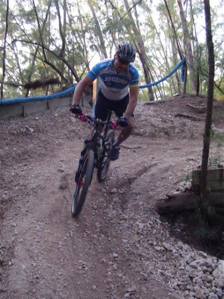 A mountain biker in a blue and white jersey rides downhill on a dirt path surrounded by trees. The biker is leaning to one side, showcasing a dynamic posture as they navigate a curve in the trail. Oleta River State Park mountain bike trail.