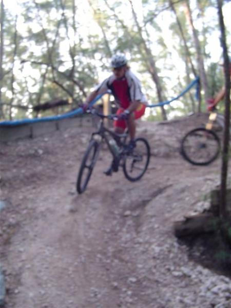 A mountain biker wearing a helmet and cycling gear jumps over a dirt mound on a forest trail, with trees in the background and another cyclist visible in the distance. Oleta River State Park mountain bike trail.
