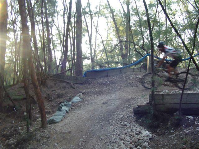A mountain biker riding over a dirt trail with a wooden ramp in a wooded area, surrounded by trees and natural sunlight filtering through the foliage. Oleta River State Park mountain bike trail.