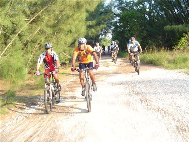 A group of cyclists riding mountain bikes along a gravel path surrounded by trees. Two riders are in the foreground, one wearing a red and white jersey and the other in an orange shirt. Several more cyclists are visible in the background, pedaling through a lush green environment. Oleta River State Park mountain bike trail.