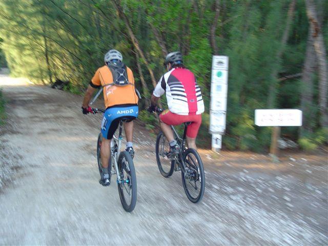 Two cyclists riding mountain bikes on a gravel path surrounded by trees. The first cyclist wears an orange jersey with a backpack, while the second cyclist is clad in a red and white jersey. A sign can be seen in the background indicating the trail. Oleta River State Park mountain bike trail.