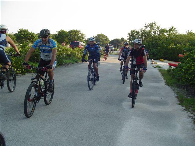 A group of four mountain bikers riding along a paved path surrounded by greenery. They are wearing cycling jerseys and helmets, with varying styles and colors. The scene captures a recreational biking activity in a natural setting on a sunny day. Oleta River State Park mountain bike trail.