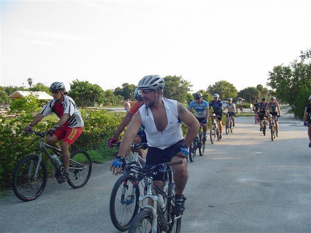 A group of mountain bikers riding on a path, surrounded by greenery and trees. Some cyclists are wearing helmets and jerseys, while one cyclist is dressed in a sleeveless shirt. The scene captures a warm, sunny day suitable for outdoor activities. Oleta River State Park mountain bike trail.